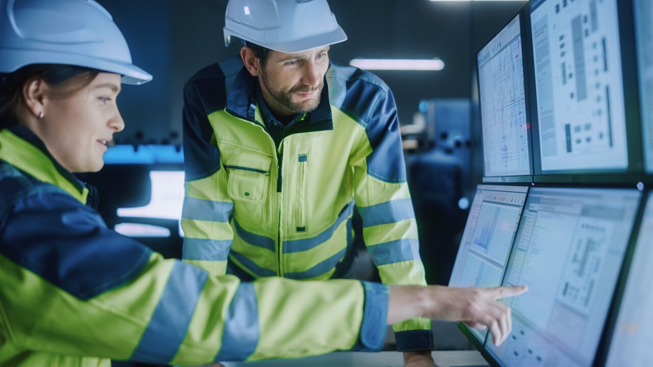 Two employees in safety gear looking at data on computer screens
